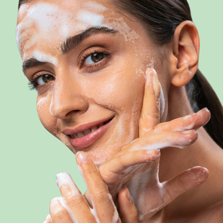 Eine lächelnde Frau trägt sanft einen schäumenden Gesichtsreiniger auf ihr Gesicht auf und vermittelt dabei ein Gefühl von Ruhe und Selbstpflege. Ihr Haar ist nach hinten gekämmt, wodurch ihre klare Haut besonders gut zur Geltung kommt.